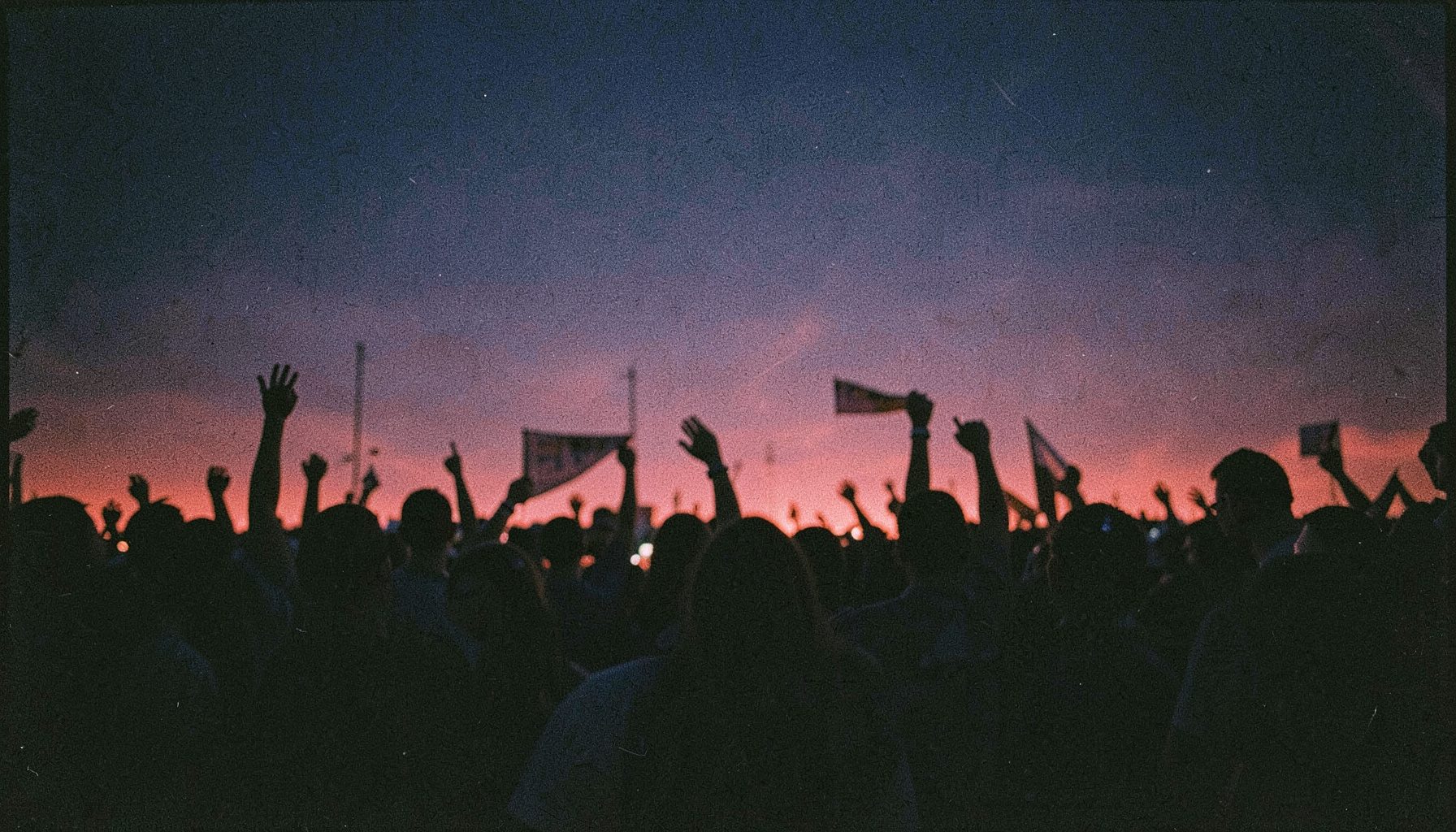 A grainy analog film photograph of a dense crowd at twilight, silhouettes of raised hands and banners against a deep navy and crimson sky, moody and defiant.