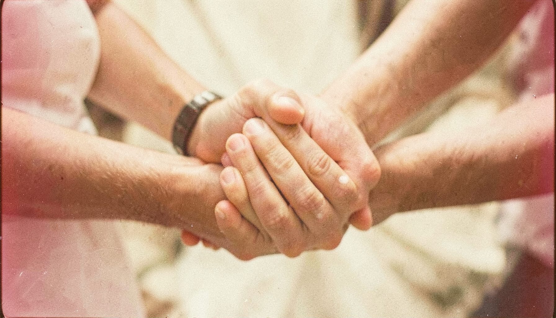 A grainy analog film photograph of hands clasped in a circle, soft natural light, cream and beige tones, hopeful and grounded.