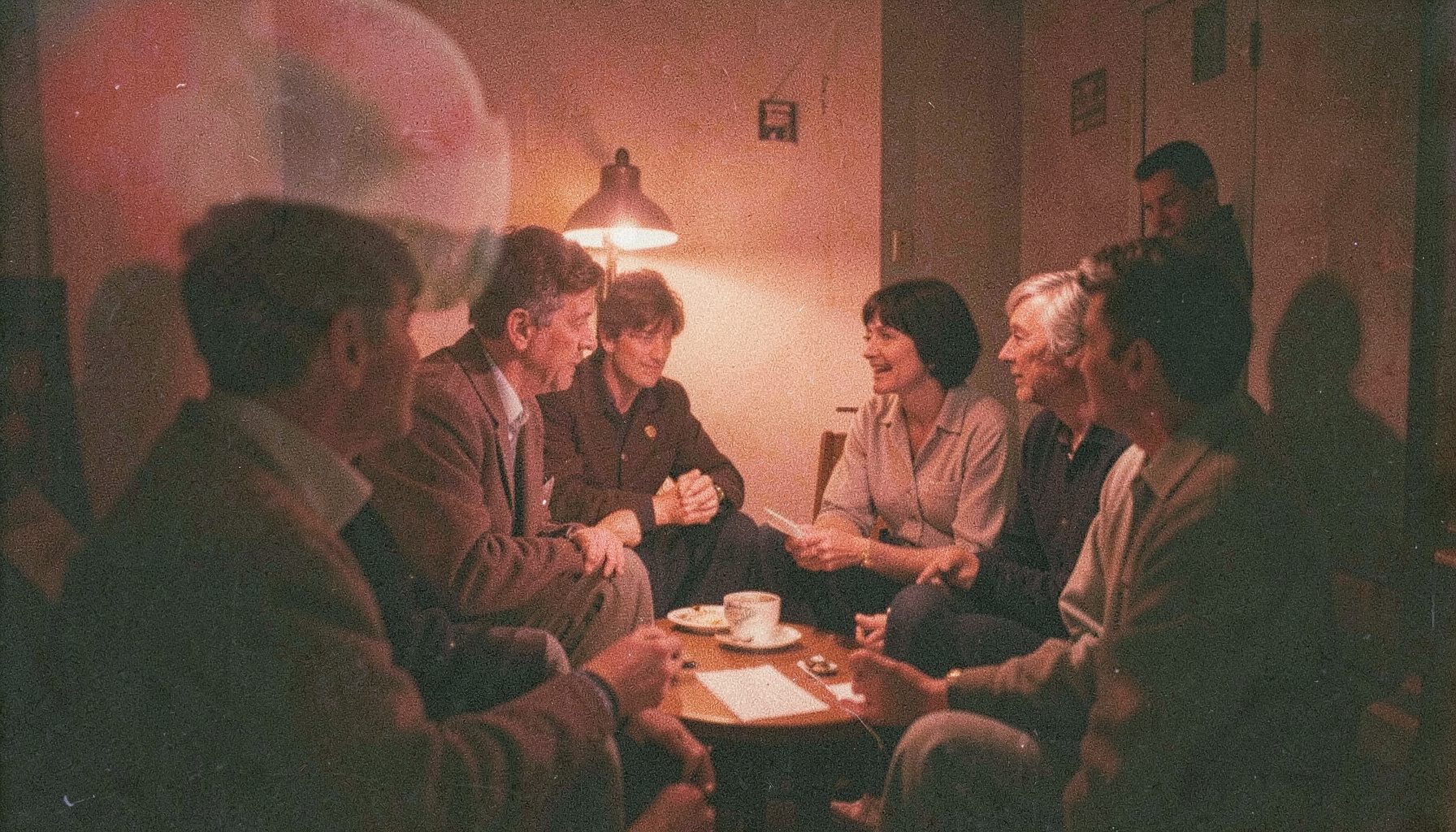 A grainy analog film photograph of a small discussion circle in a dim room, warm lamp light, intimate and thoughtful atmosphere.