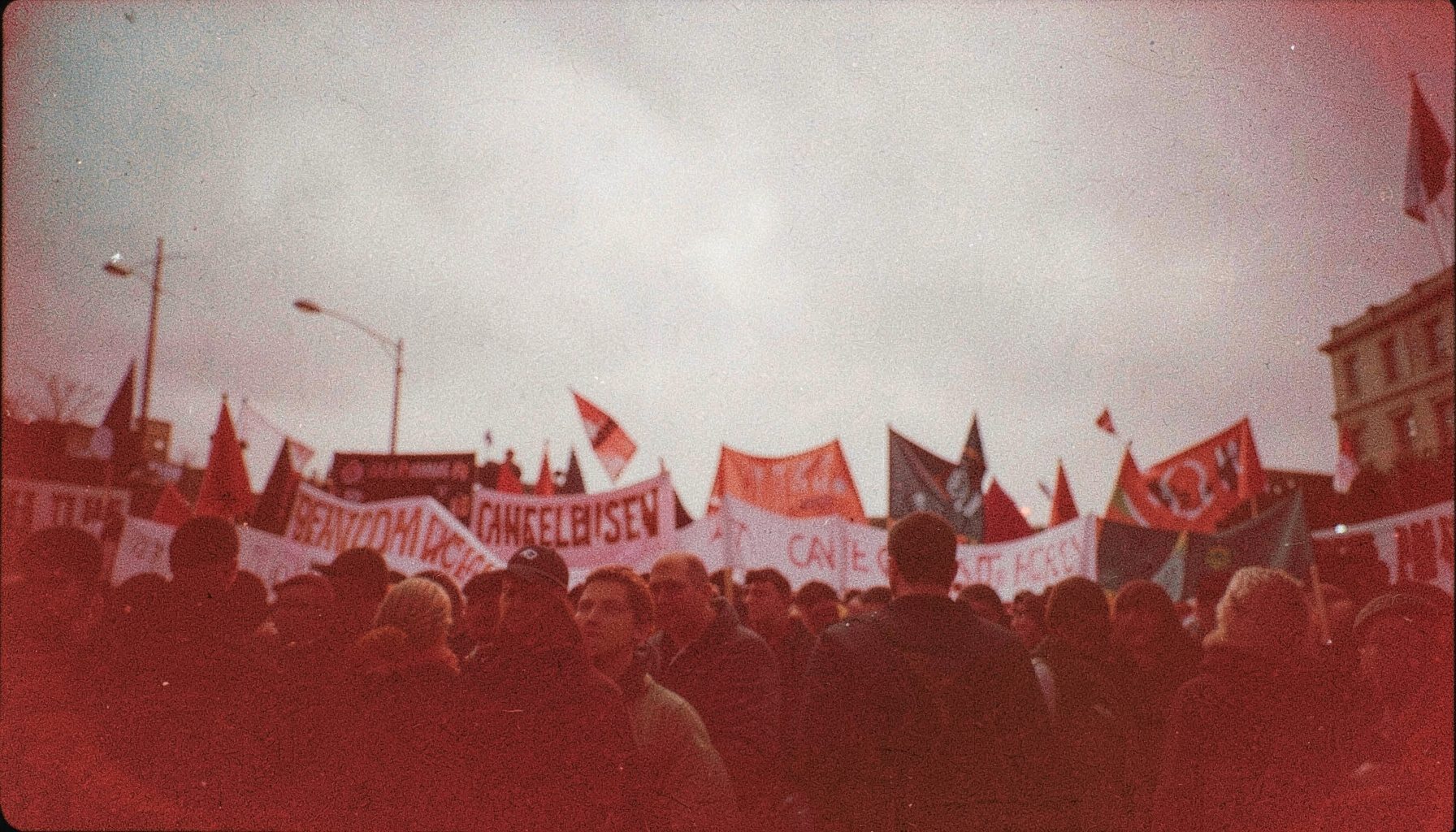 A grainy analog film photograph of a protest march, banners silhouetted against an overcast sky, deep reds and muted grays.