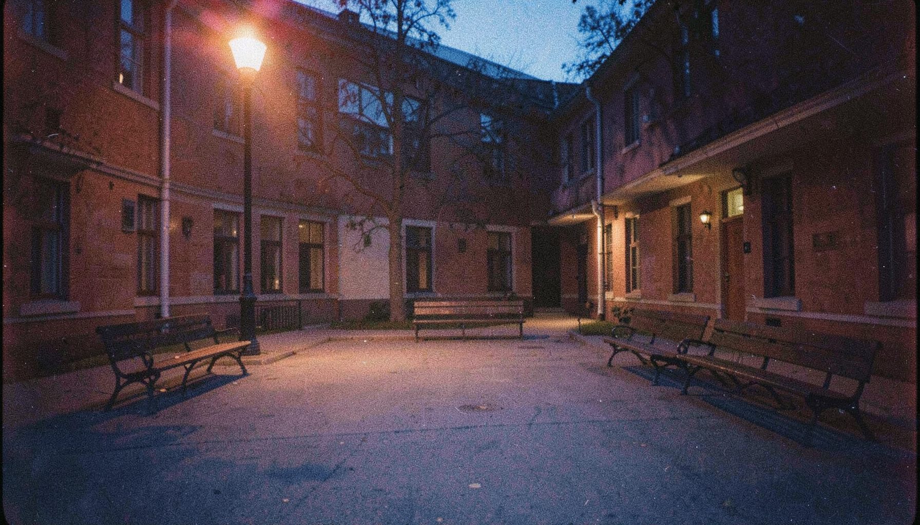 A grainy analog film photograph of a quiet campus courtyard at dusk, empty benches under warm streetlight, navy blue and amber tones, contemplative.