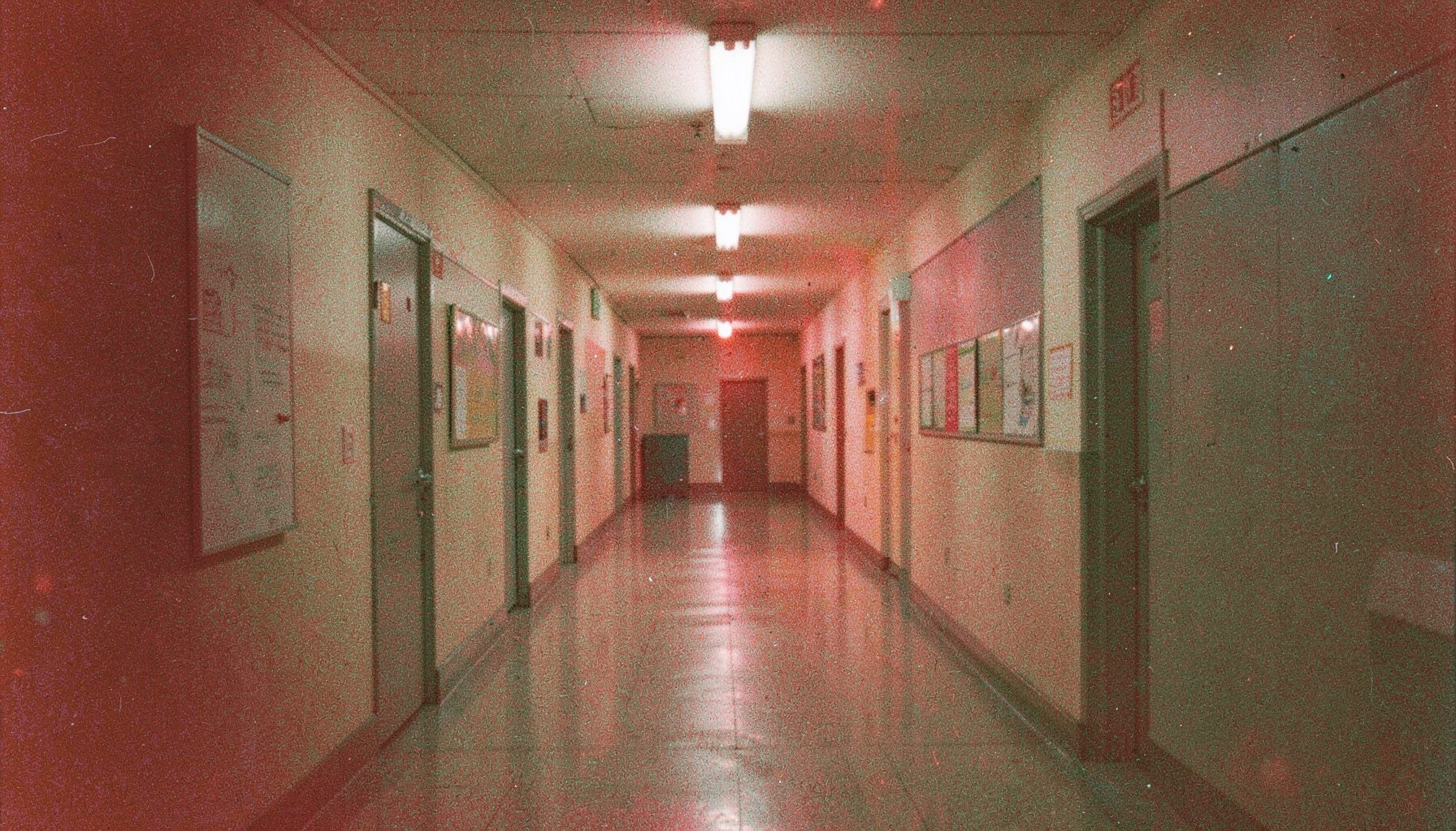 A grainy analog film photograph of a school corridor after hours, fluorescent light casting long shadows, quiet and uneasy.