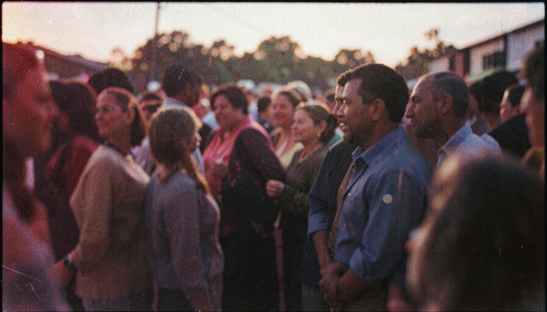 A grainy analog film photograph of a crowded community gathering at dusk, warm amber and deep indigo tones, soft blur at the edges, people standing close together in quiet solidarity.