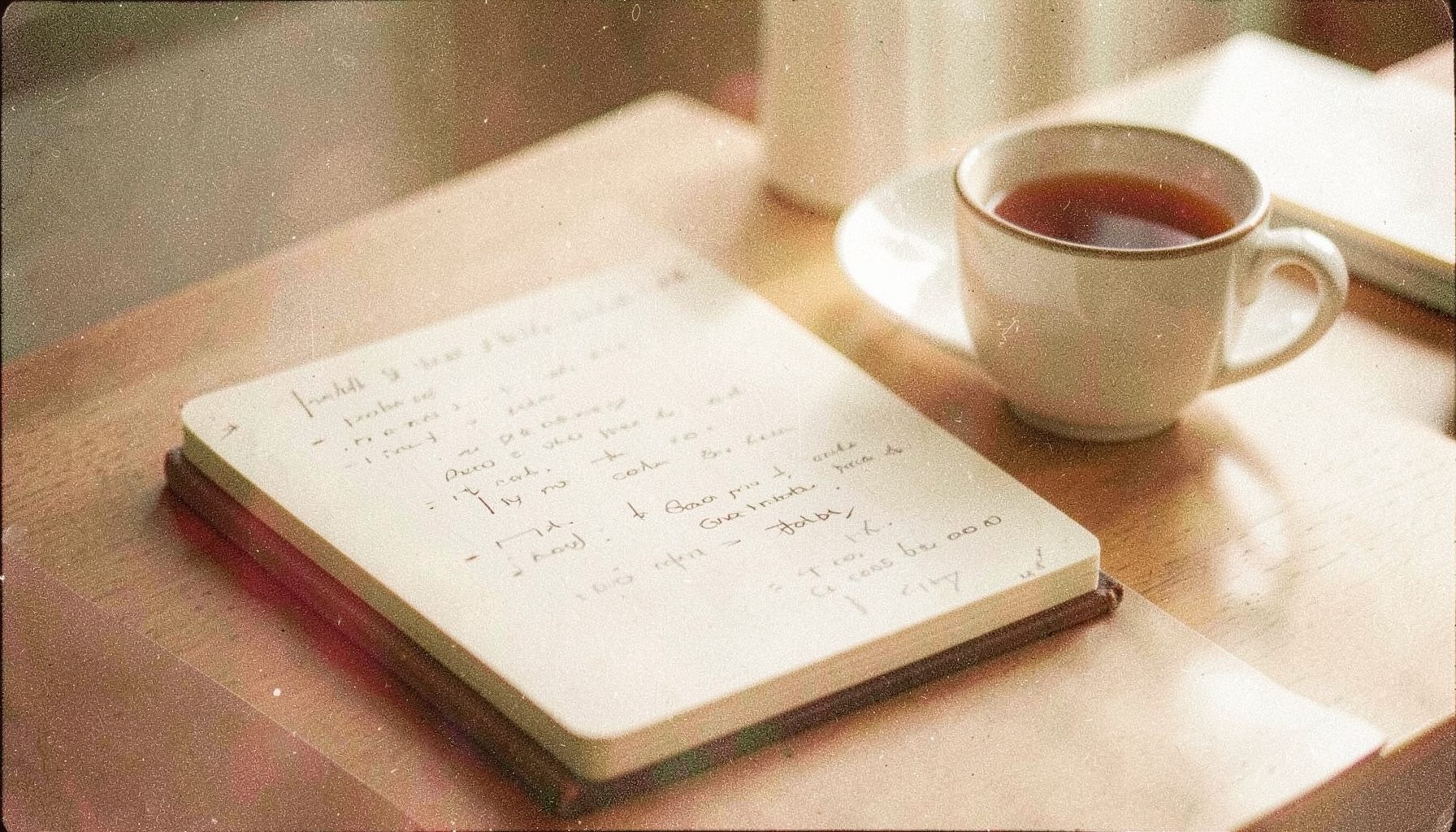 An analog film photograph with visible grain, a single open notebook with handwritten notes beside a cup of tea on a sunlit desk, soft beige and muted gold tones.