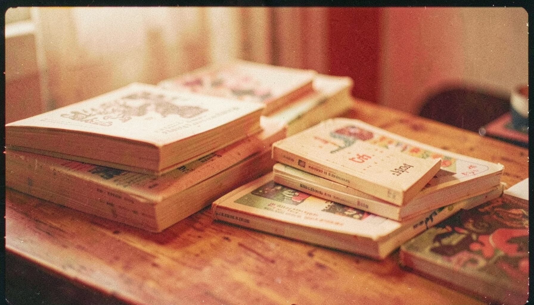 A softly lit analog film photograph with visible grain, showing a stack of well-worn paperback books and zines spread across a wooden table, warm amber and cream tones, quiet and intimate atmosphere.