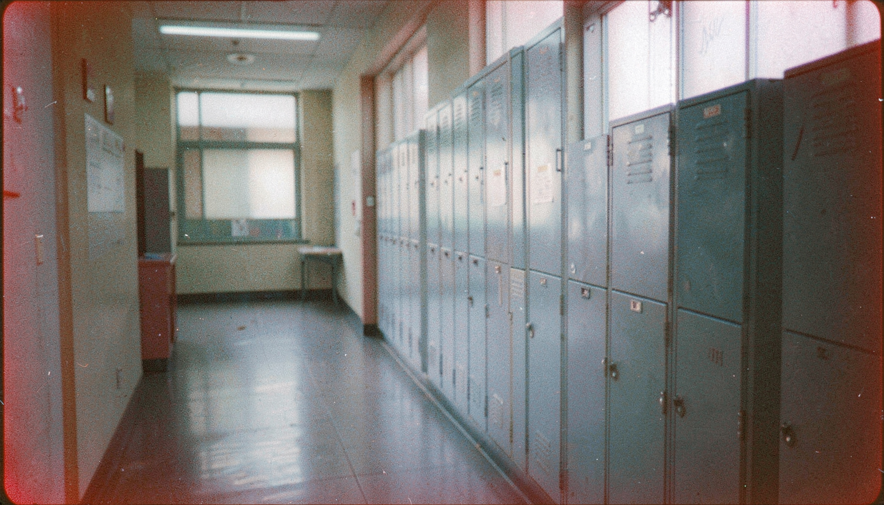 An analog film photograph with visible grain, a quiet school corridor with lockers lining one wall, diffused afternoon light through frosted windows, muted blues and pale grays.