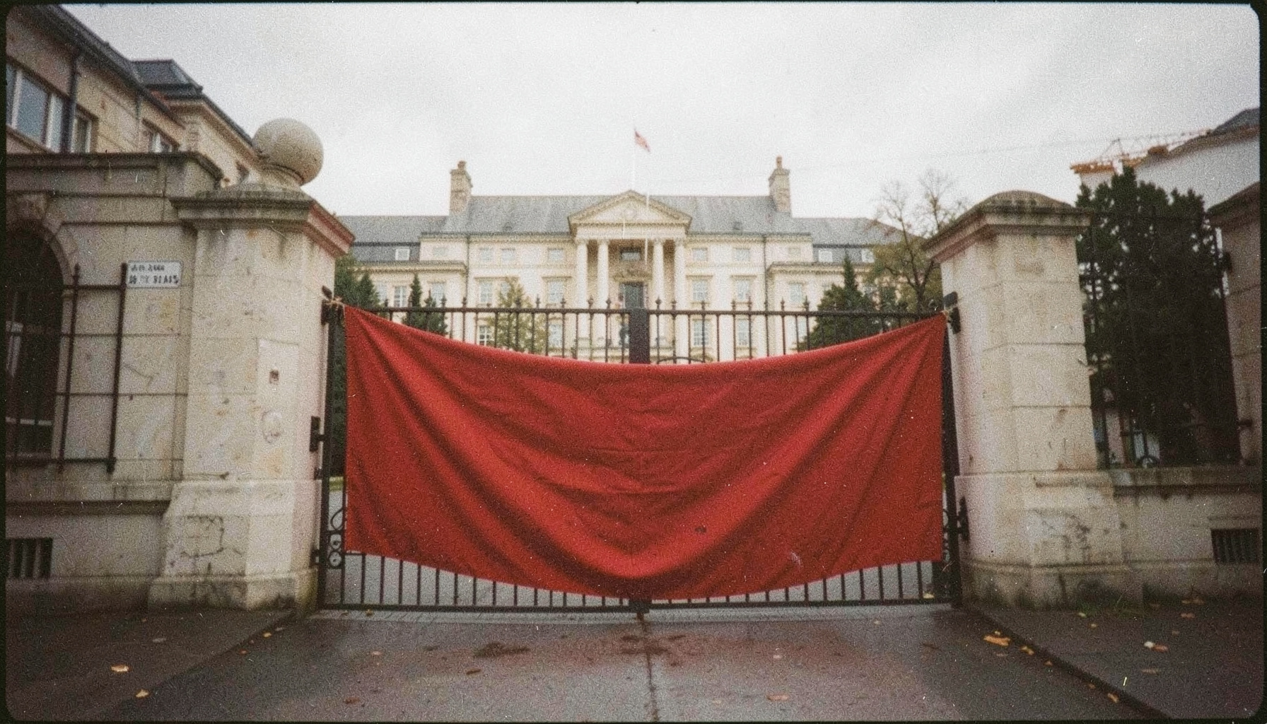 An analog film photograph with visible grain, a protest banner draped across a campus gate, deep red fabric against pale stone, overcast sky, muted and earnest tones.