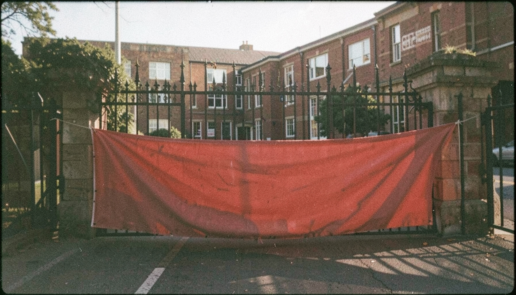 A grainy analog film photograph of a protest banner draped across a university campus gate, deep reds and muted earth tones, soft morning light casting long shadows.