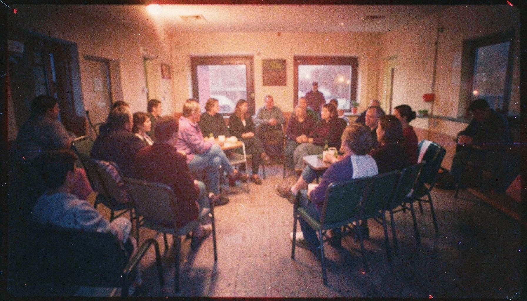 A grainy analog film photograph of a warmly lit community gathering space at dusk, with soft amber and deep indigo tones, empty chairs arranged in a loose circle suggesting conversation and solidarity.